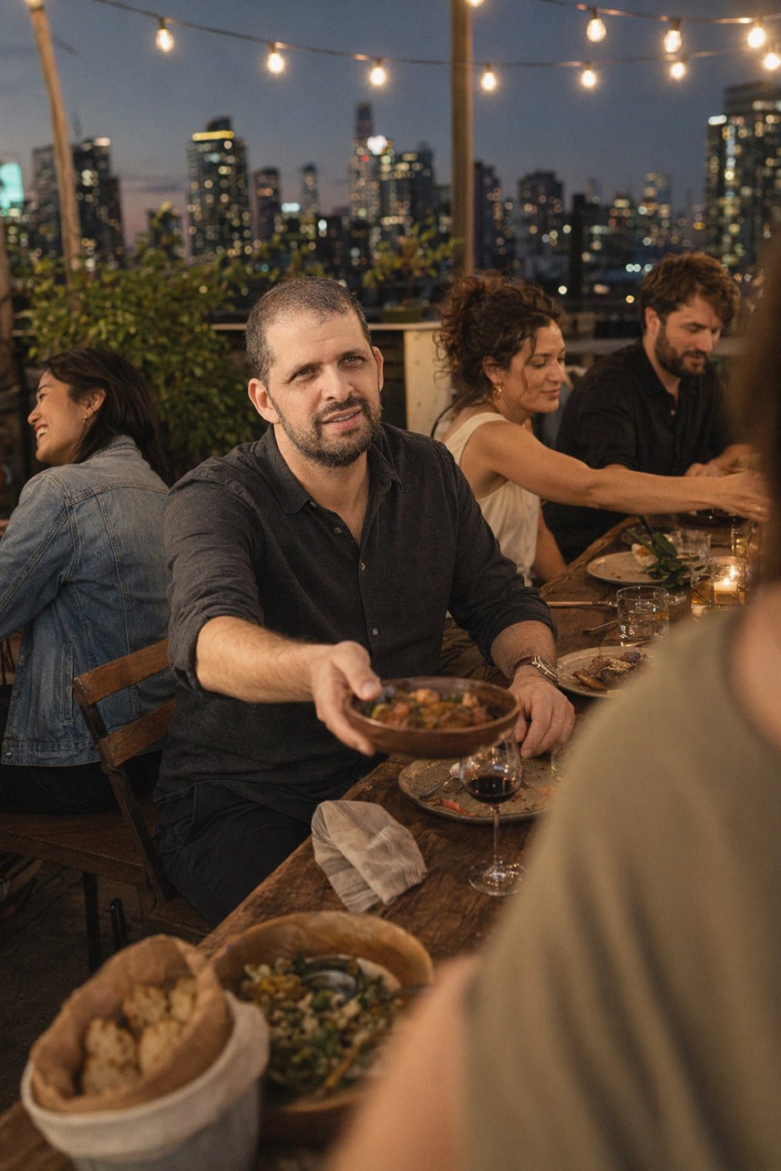 Man passing a dish at a rooftop dinner table under string lights