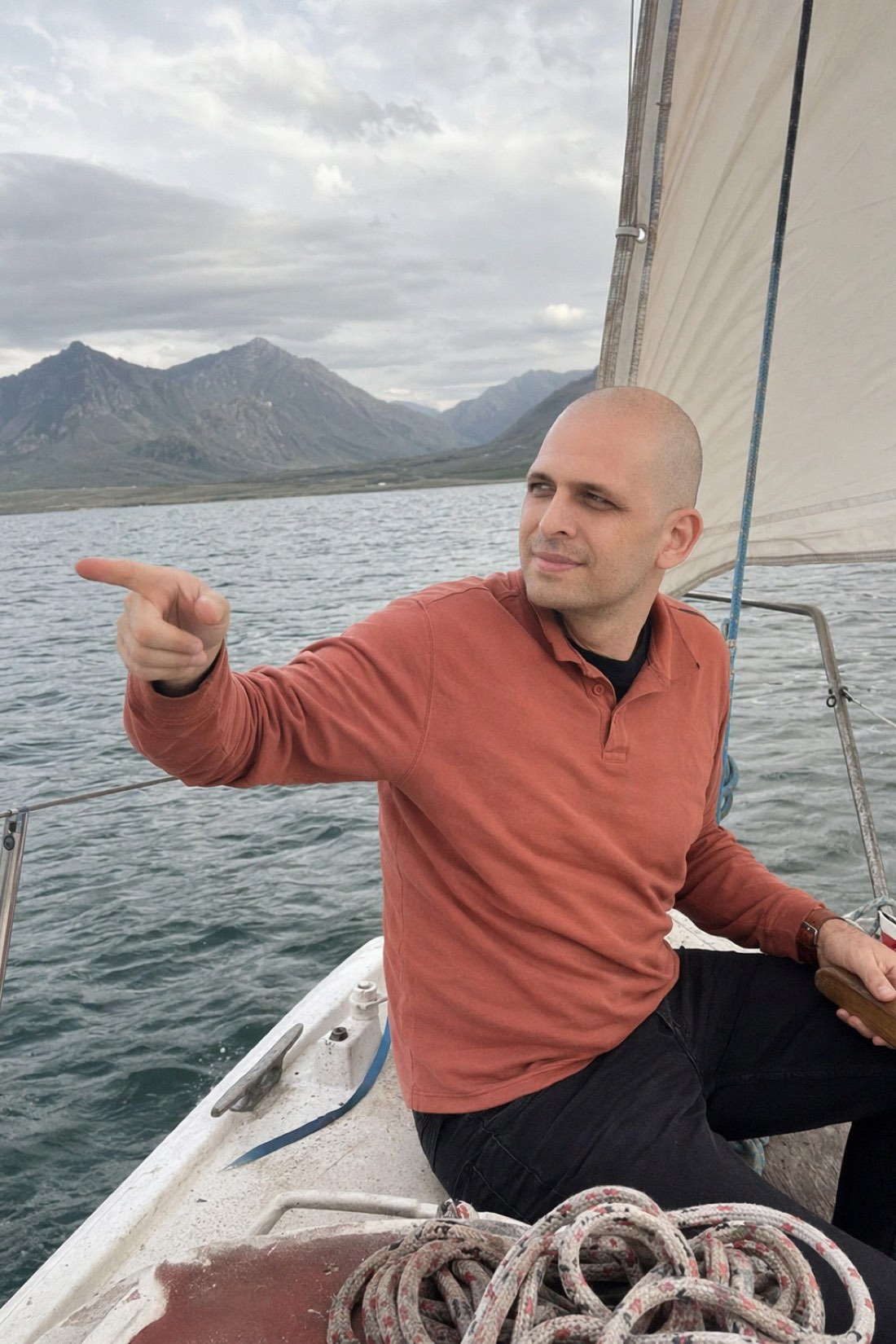 Man pointing from a sailboat with mountains and a lake behind him, a realistic AI travel photo